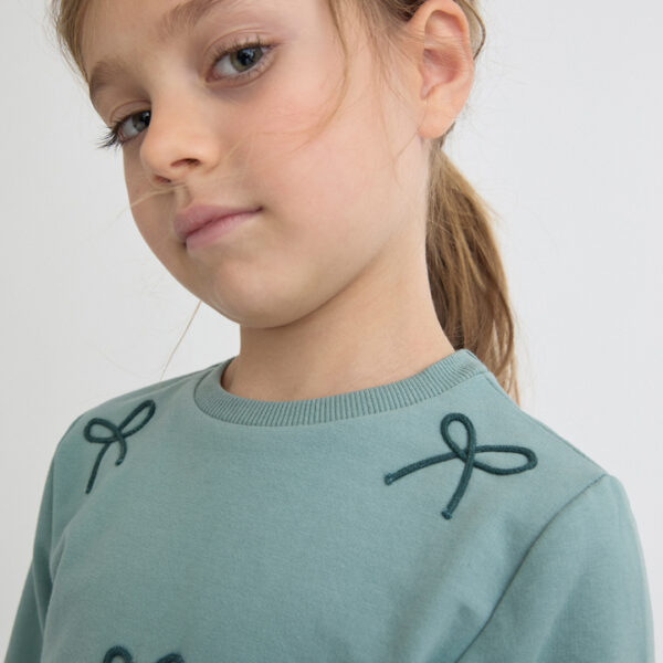 Sudadera azul verdoso con apliques de lazos bordados en el frontal y volante de tul en el bajo.