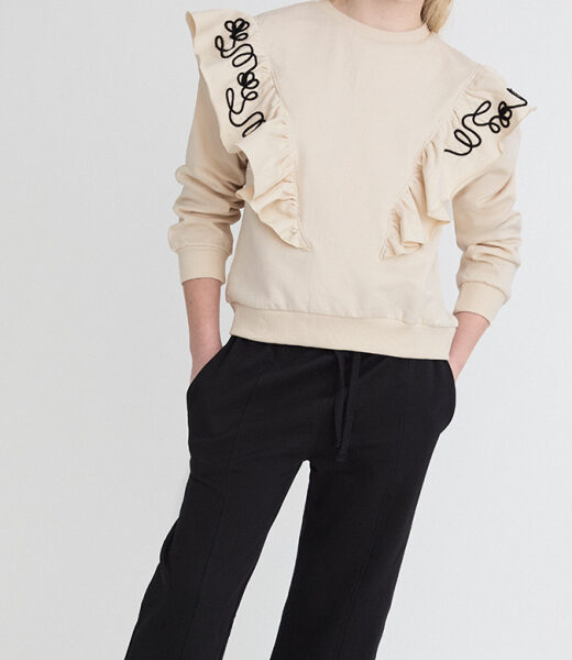 Conjunto con sudadera beige de felpa suave con volantes y bordado decorativo en hombros. Pantalón negro de punto elástico, cálido y confortable.